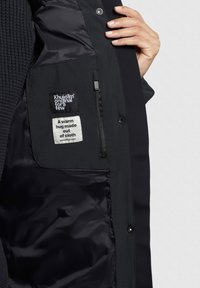 Chaqueta negra con un forro interior suave; presenta un tejido texturizado, etiquetas con texto impreso y un bolsillo con cremallera en el interior.