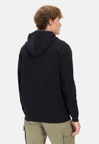 Schwarzer Kapuzenpullover mit strukturiertem Rippdesign, langen Ärmeln und lässiger Passform. Besitzt ein rückseitiges Nahtdetail und wird mit hellen Hosen kombiniert.