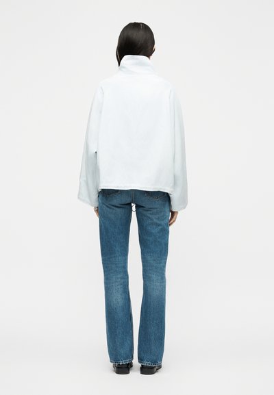 Η γυναίκα στέκεται γυρισμένη με την πλάτη, φορώντας ένα λευκό μπουφάν με ψηλό γιακά, μπλε τζιν με ίσια γραμμή και μαύρα παπούτσια σε απλό φόντο.