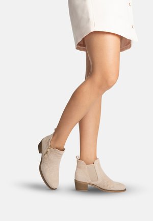 Benen met lichtbeige geperforeerde enkellaarsjes met kleine blokhakken en ritssluitingen aan de zijkant, gecombineerd met een lichtbeige rok.