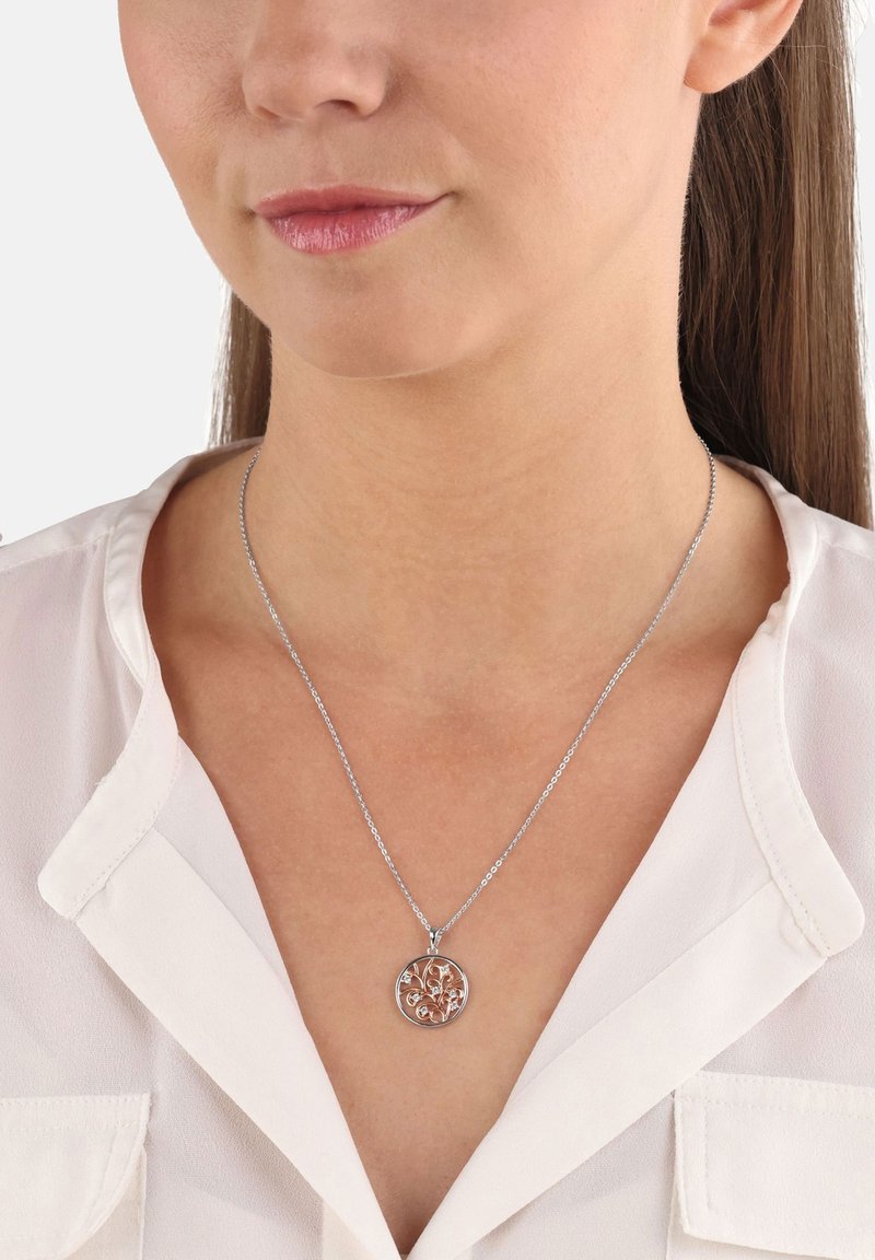 Donna che indossa una collana con pendente d'argento con un design floreale su una catena delicata sopra una blusa bianca.