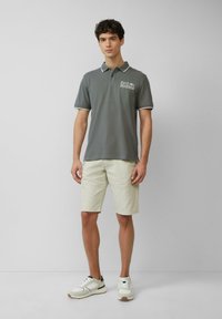 Grijze polo met witte accenten, voorzien van een Ford Mustang-logo. Gekozen in combinatie met lichtbeige shorts en witte sneakers met marineblauwe accenten.