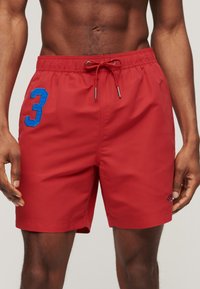 Männlicher Torso trägt rote Badeshorts mit einer blauen Nummer 3 am linken Oberschenkel und einem Kordelzug am Bund, steht vor einem einfarbigen Hintergrund.