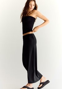 Un top tube noir ajusté associé à une jupe maxi fluide taille haute en tissu lisse et extensible. Porté avec des tongs noires.