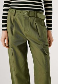 Persoon in olijfgroene cargobroek met klepzakken en stoffen riem, gecombineerd met een zwart-wit gestreepte shirt met lange mouwen.