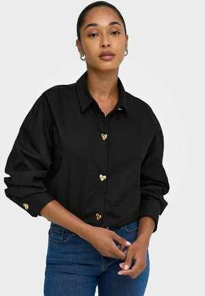 Femme portant une chemise noire avec des boutons en forme de cœur dorés et un jean bleu, debout devant un fond clair uni.