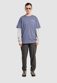 Fioletowa,oversized koszulka nałożona na szary, długi rękaw, w połączeniu z ciemnoszarymi spodniami i beżowymi butami trekkingowymi. Kątowe okulary przeciwsłoneczne dopełniają całość.