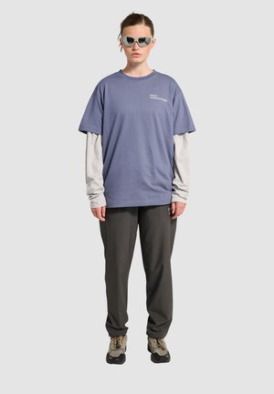 Lila oversized t-shirt lagerad över en grå långärmad tröja, tillsammans med mörkgrå byxor och beige vandringsskor. Vinklade solglasögon fulländar looken.