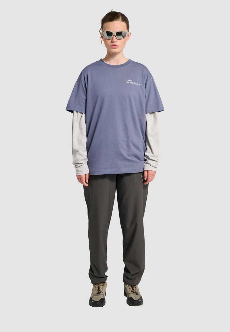 Fioletowa,oversized koszulka nałożona na szary, długi rękaw, w połączeniu z ciemnoszarymi spodniami i beżowymi butami trekkingowymi. Kątowe okulary przeciwsłoneczne dopełniają całość.