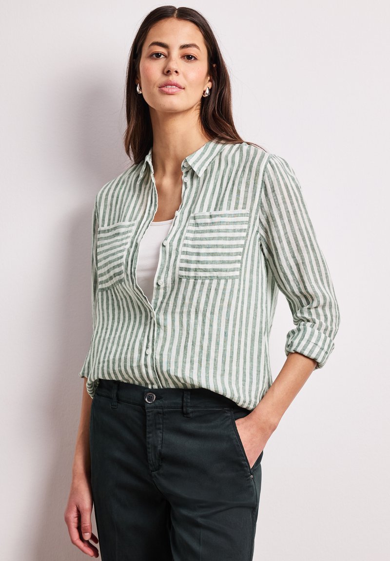 Femme portant une chemise à boutons à rayures verticales vertes et blanches sur un haut blanc avec un pantalon noir, debout avec une main dans la poche.