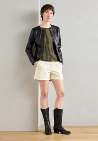 Black faux leather jacket with a fitted design, olive green tie-dye top, cream shorts, and black knee-high boots with pointed toes.