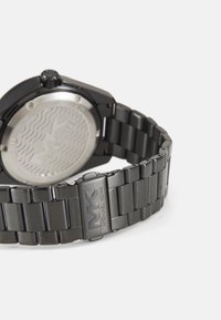 Reloj de metal negro con un diseño de pulsera elegante. La caja cuadrada presenta un respaldo plateado texturizado con un patrón ondulado y detalle del logotipo.