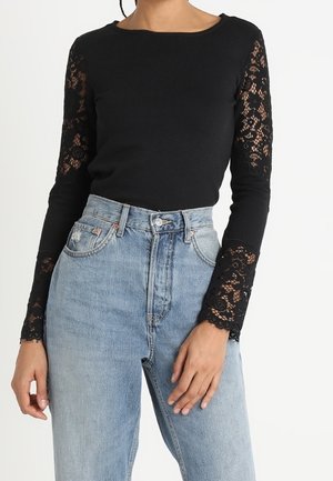 Kobieta w czarnej bluzce z długim rękawem z koronkowymi rękawami i wysokim stanem jasnoniebieskich jeansów, stojąca z jedną ręką przy kieszeni.