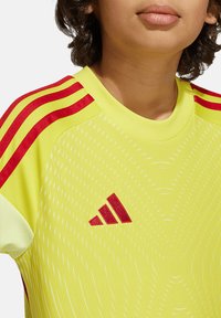 T-shirt de sport jaune avec des rayures rouges sur les manches, présentant un motif texturé et un logo Adidas rouge sur le devant.