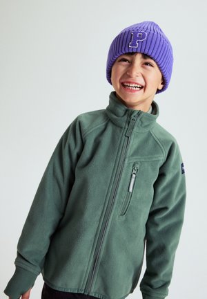Smilende barn iført en grøn fleecejakke med lynlås og en lilla strikhue med bogstavet "P" på, stående mod en ensfarvet baggrund.