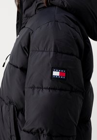Veste matelassée noire avec capuche, présentant des surpiqûres horizontales, une poche zippée latérale et un patch logo rectangulaire aux accents bleu, blanc et rouge.