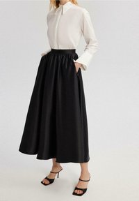 Falda negra de satén hasta el tobillo con cintura elástica y bolsillos laterales, combinada con una blusa blanca de cuello y tacones negros con tiras.