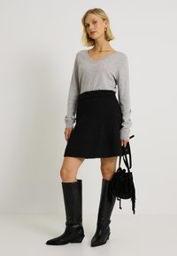 Pull gris à col en V, jupe en tricot noire, bottes en cuir noir jusqu'au genou. Sac à main noir avec des détails à franges. Silhouette simple et ajustée.