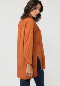Blusa de manga larga en naranja óxido, con un corte relajado, aberturas laterales, puños abotonados y un yugo trasero plisado en tejido ligero.