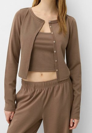 Femme portant un cardigan court côtelé marron sur un débardeur assorti et un pantalon à taille élastique, montrant son ventre et ses longs cheveux.