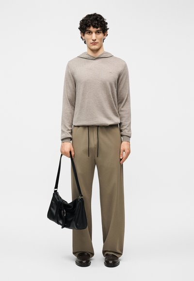 Νέος άνδρας όρθιος, φορώντας μπεζ φούτερ με κουκούλα και καφέ παντελόνι φαρδύ, κρατώντας μαύρη δερμάτινη τσάντα στο δεξί χέρι, σε λευκό φόντο.