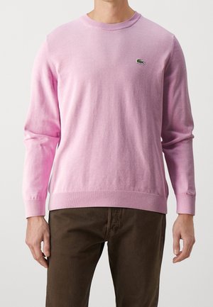 Homme portant un pull rose clair avec un petit logo crocodile vert sur la poitrine gauche et un pantalon marron foncé.