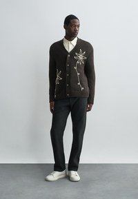 Barna kötött kardigán v-nyakkal, krémszínű virágmintával, sötét nadrággal és fehér sportcipővel párosítva. Egyszerű háttér.