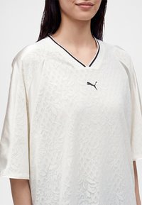 Γυναίκα φοράει λευκό αθλητικό πουκάμισο Puma με δαντελωτό σχέδιο, μαύρο λογότυπο Puma και V-λαιμόκοψη με μαύρη επένδυση.