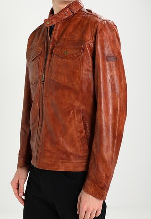 Brun skindjakke med lynlås foran, to brystlommer med klap og knapper samt krave med trykknapslukning båret af en person i sorte bukser.