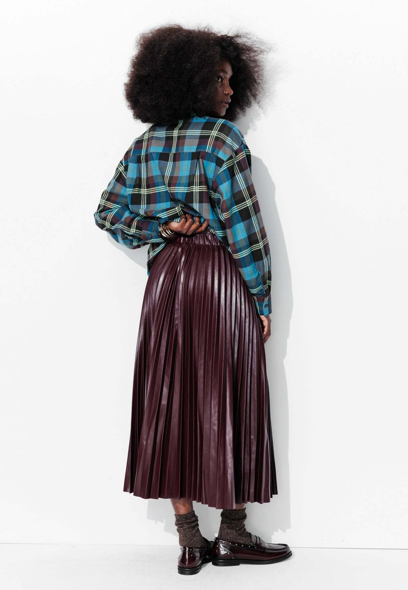 Chemise à carreaux bleue et noire, associée à une jupe plissée en simili cuir bordeaux. Chaussures noires brillantes et chaussettes marron texturées.