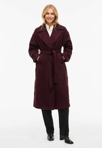 Burgundy wrap coat with a tie belt, wide lapels, and side pockets. Soft textured fabric and knee-length, paired with black pants and boots.