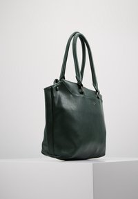 Ciemnozielona skórzana torba tote z podwójnymi uchwytami, wyeksponowana w pozycji pionowej na białym postumencie na tle neutralnym.