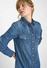 Chemise en denim dotée de manches longues, d'un col boutonné, de deux poches poitrine et d'une couleur bleu clair avec une texture subtile et des détails de couture.