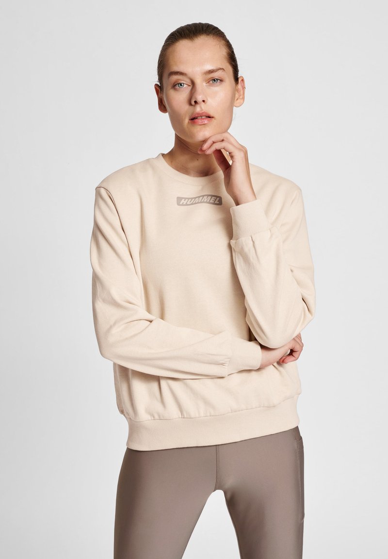 Beige sweatshirt med rund hals, långa ärmar och ribbbade ärmslut. Har en subtil logotyp på bröstet. Kombineras med bruna leggings.