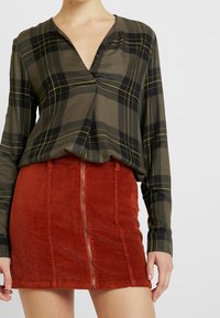 Gestreifte Bluse mit langen Ärmeln in olivgrün, verziert mit schwarzen und gelben Linien, kombiniert mit einem rostorangefarbenen Cord-Rock mit Mini-Länge und einem Vorderreißverschluss.