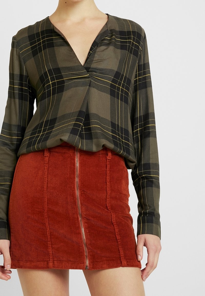 Gestreifte Bluse mit langen Ärmeln in olivgrün, verziert mit schwarzen und gelben Linien, kombiniert mit einem rostorangefarbenen Cord-Rock mit Mini-Länge und einem Vorderreißverschluss.