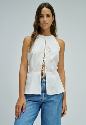 Mujer con cabello largo y castaño que lleva pendientes de aro dorados, una camiseta blanca sin mangas con lazos delanteros y vaqueros azules.