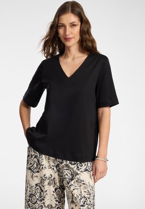 Frau mit gewelltem braunem Haar, die ein schwarzes V-Ausschnitt-Kurzarmtop und cremefarbene weite Hosen mit schwarzem Blumenmuster trägt.