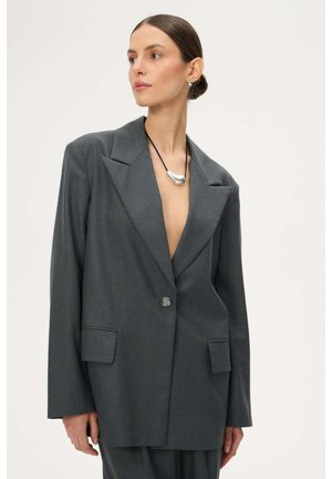 Kobieta ubrana w oversize'ową, ciemnoszarą marynarkę i pasujące spodnie, ozdobiona srebrnym naszyjnikiem z zawieszką i kolczykami wkrętami, pozująca na tle jednolitego tła.