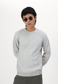 Pull en maille côtelée gris avec un col rond et des manches longues, fabriqué à partir d'un matériau en tricot texturé. Associé à des lunettes de soleil noires rondes.