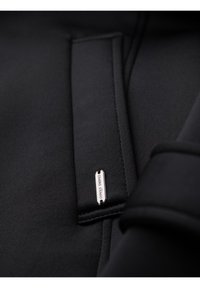 Gros plan sur le tissu d'un sweat à capuche noir montrant un cordon de serrage avec une petite étiquette métallique gravée "OMBRE EXIMIO".