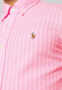 Ljusrosa skjorta med knappar som har subtila vertikala ränder, en spetsig krage och en broderad brun polo-ponny-logotyp på bröstet.