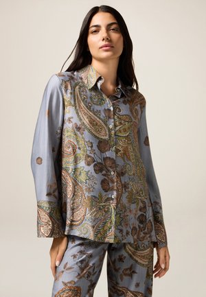 Vrouw met lang donker haar, gekleed in een bijpassende lichtblauwe paisley en bloemenpatroon knopenoverhemd en broek, staande tegen een neutrale achtergrond.