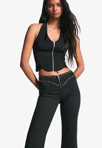 Crni skrojeni halter top s prednjim zatvaračem i prilagođenim dizajnom, uparen s crnim hlačama visokog struka s pojasom ukrašenim zakovicama i zatvaračem.