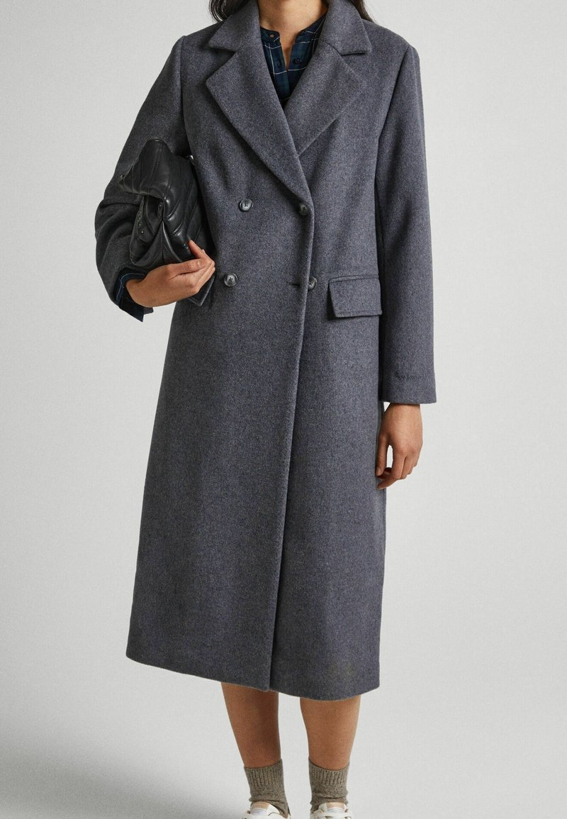 Abrigo largo de mezcla de lana gris con cuello de muesca, botones de doble botonadura, bolsillos laterales y un corte relajado. El modelo sostiene un bolso negro.