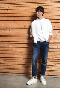 Camisa blanca de botones sobre una camiseta blanca, jeans azules y zapatillas blancas. Posado contra una pared de madera texturizada con tablones horizontales.