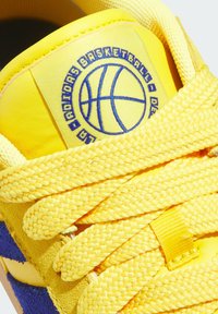 Langue de sneaker jaune avec logo de basketball bleu et texte, lacets épaisses tressés jaunes et détail en suède bleu sur le côté.
