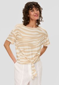 Beige T-Shirt mit kurzen Ärmeln und einem wellenförmigen weißen Muster, mit einem geknoteten Detail an der Taille, aus leichtem Stoff. Getragen mit weißen Hosen.