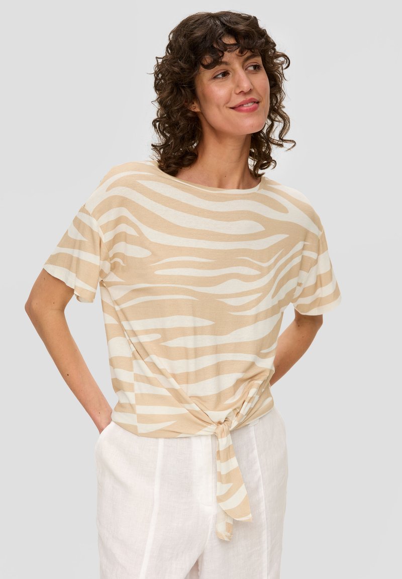 Beige T-Shirt mit kurzen Ärmeln und einem wellenförmigen weißen Muster, mit einem geknoteten Detail an der Taille, aus leichtem Stoff. Getragen mit weißen Hosen.