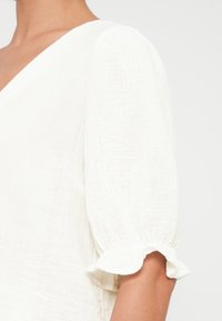 Λευκό πουκάμισο με βαθύ V-λαιμό και κοντά φουσκωτά μανίκια. Το ύφασμα έχει πτυχιαστή υφή και βολάν στις άκρες των μανικιών.